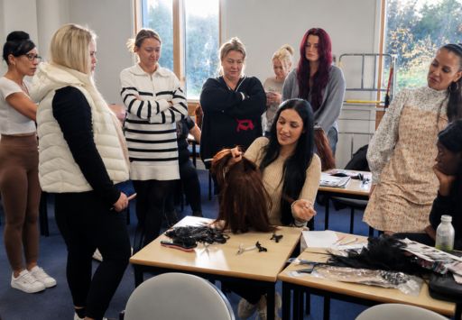 Small group hair extension training session in a UK academy with a tutor demonstrating technique on a mannequin head while students observe and practice using professional tools.