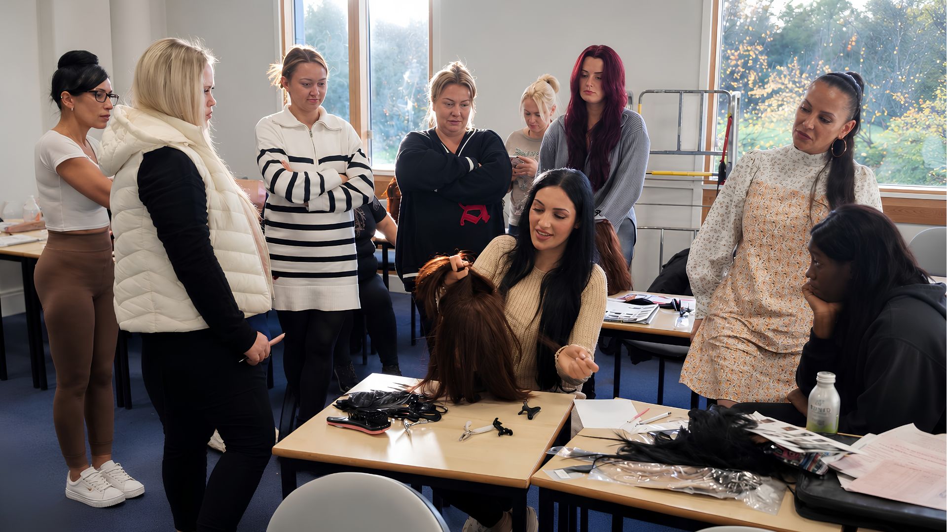 Small group hair extension training session in a UK academy with a tutor demonstrating technique on a mannequin head while students observe and practice using professional tools .